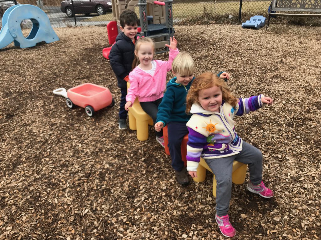 Preschool students making a pretend train with chairs on the playground