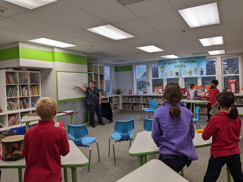 A music teacher pointing at a whiteboard with rhythms on it while students play percussion instruments