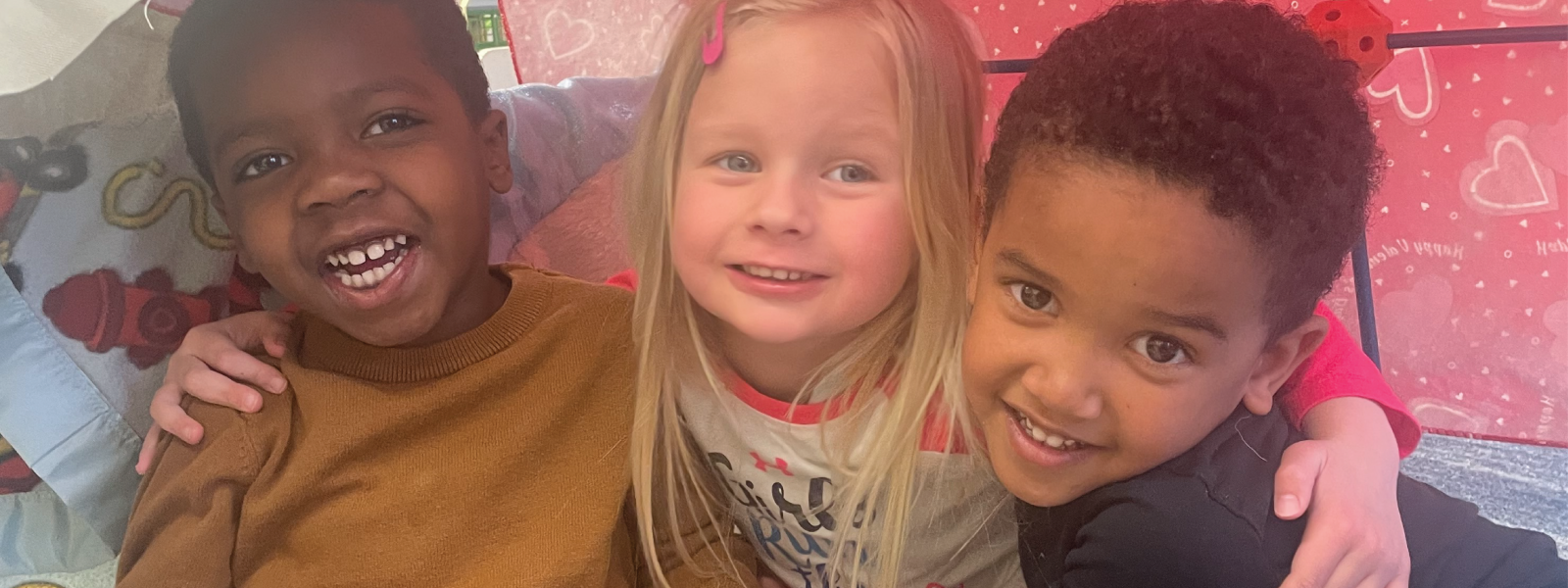 three smiling preschool friends sitting in a blanket fort with their arms around each other