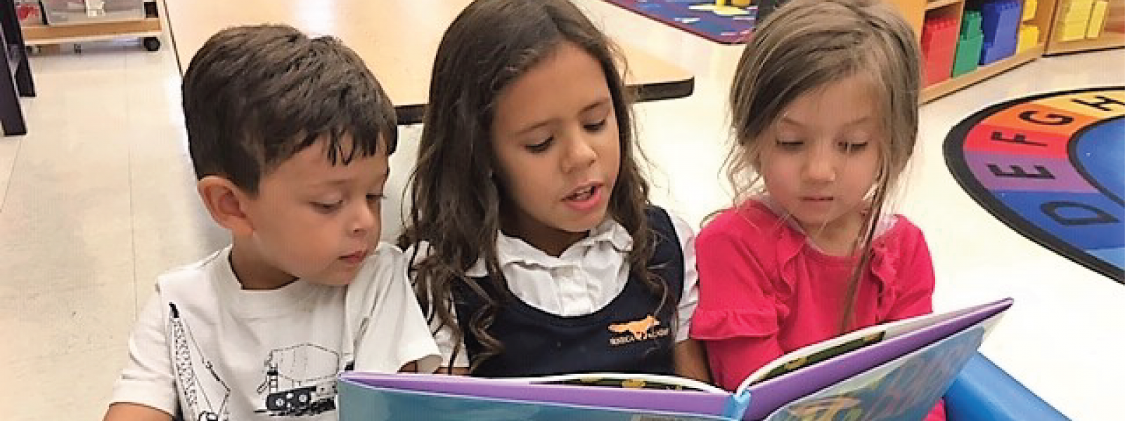 an elementary school girl sitting in between two preschoolers, reading a book aloud to them
